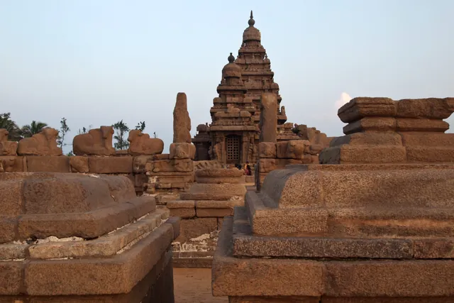 A photograph of the Shore Temple at Mamallapuram at dusk, its stepped granite tower rising behind low weathered walls and carved blocks.