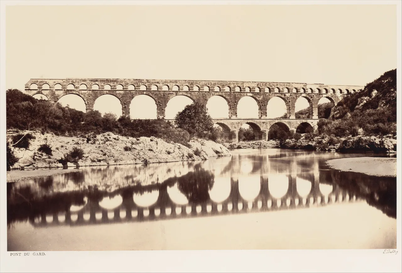 A sepia photograph of the Roman aqueduct bridge of the Pont du Gard, three tiers of stone arches stacked one above another, reflected in the still river below.
