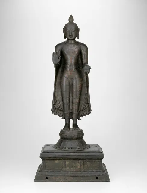 A bronze standing Buddha from Nagapattinam, one hand raised and the other lowered in the gesture of giving, his robe falling in fine folds over a lotus and stepped base.