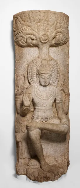 A tall granite relief of Shiva seated under a tree as the teacher, one leg folded, with a calm face and raised hand.