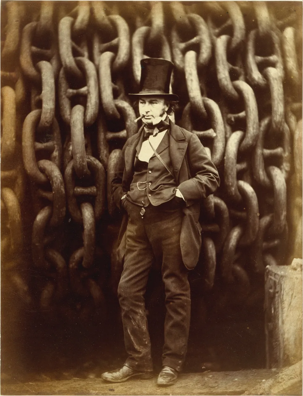 A sepia photograph of the engineer Brunel in a tall hat, hands in pockets, standing in front of a wall of enormous iron chain links at the shipyard.