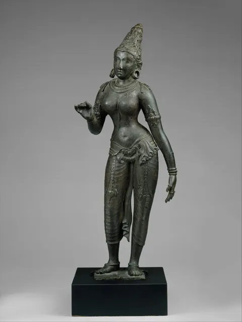 A slender bronze of the goddess Parvati standing in a gentle sway, one hand raised, wearing a tall crown and a finely incised lower garment.