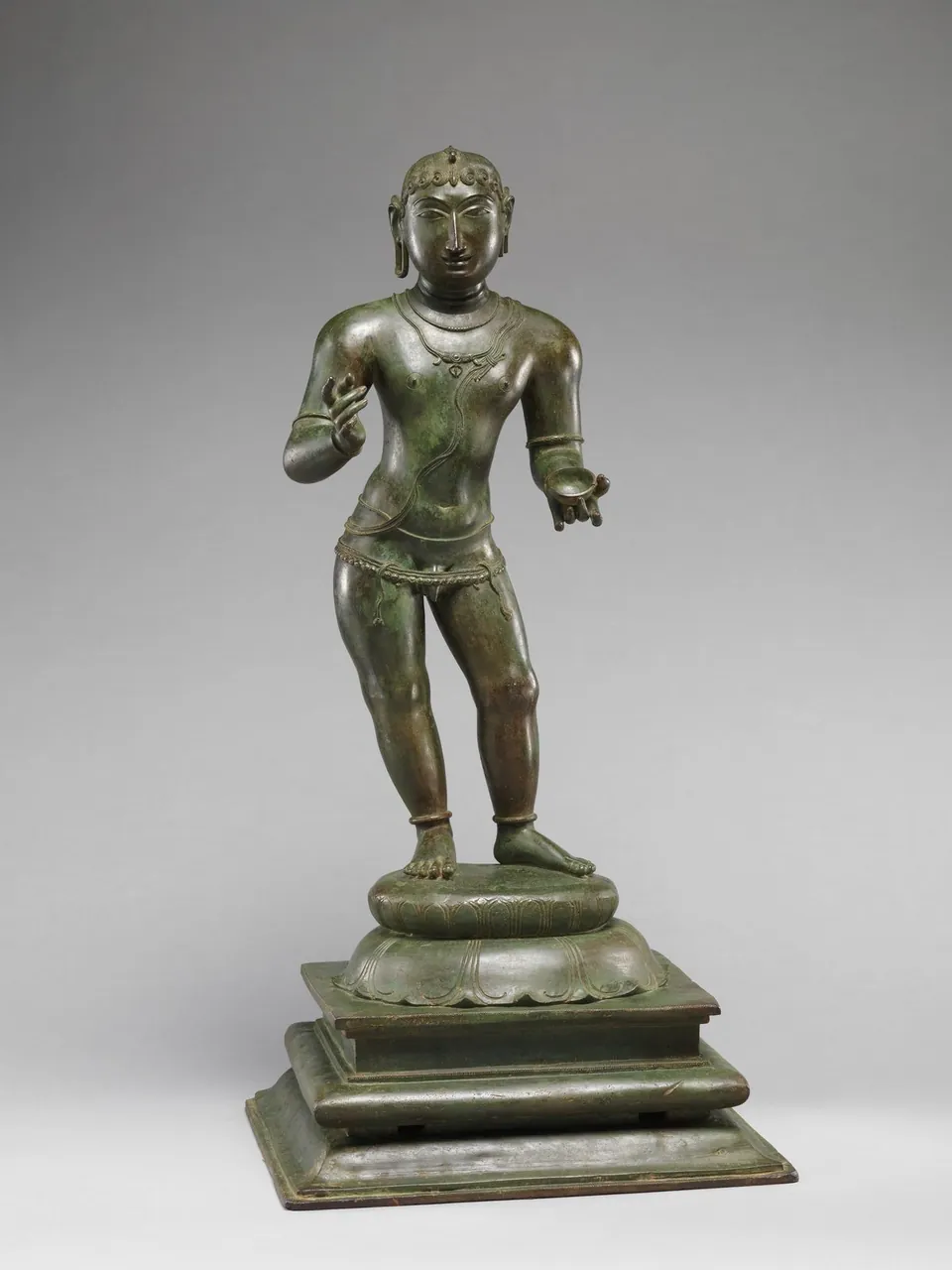A bronze statue of the child saint Sambandar standing on a lotus base, one hand raised and pointing upward.