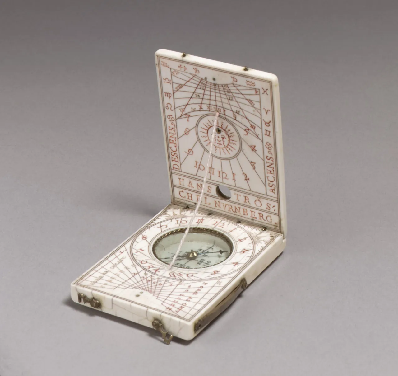 A small folding ivory sundial opened at a right angle, its two leaves engraved with hour lines in red and black, a compass set into the lower leaf.